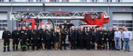 Uroczyste pożegnanie strażaka odchodzącego na zaopatrzenie emerytalne - wspólne zdjęcie funkcjonariuszy oraz gości. W tle drabina pożarnicza.