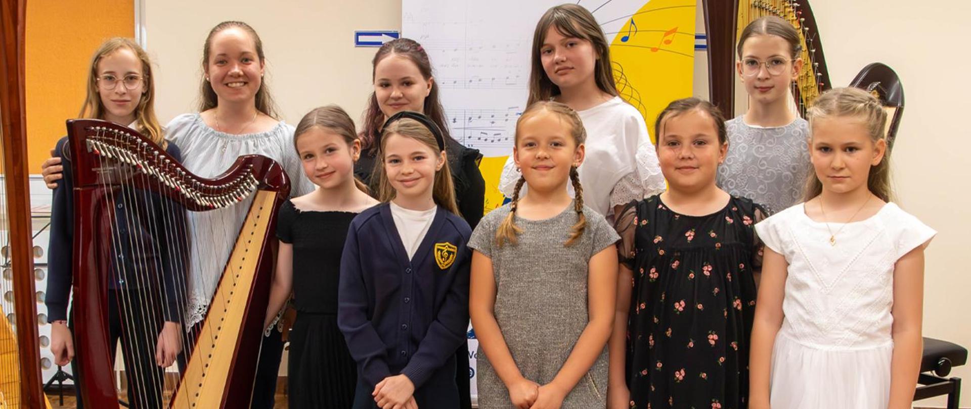 Na zdjęciu nauczycielka i 9 dziewczynek stoją w auli obok harf, za nimi stoi baner szkolny.