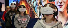Na krzesłach siedzi grupa kolorowo ubranych dzieci, na głowach mają gogle rzeczywistości wirtualnej z napisem Innowacyjna Historia, za nimi kolorowa ściana.