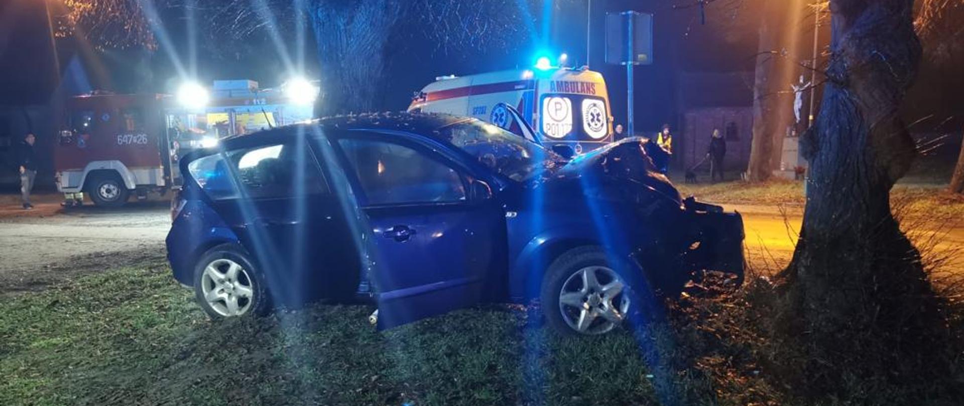 Na zdjęciu widać uszkodzony granatowy samochód osobowy, w tle karetka pogotowia oraz samochód strażacki oświetlający teren działań.