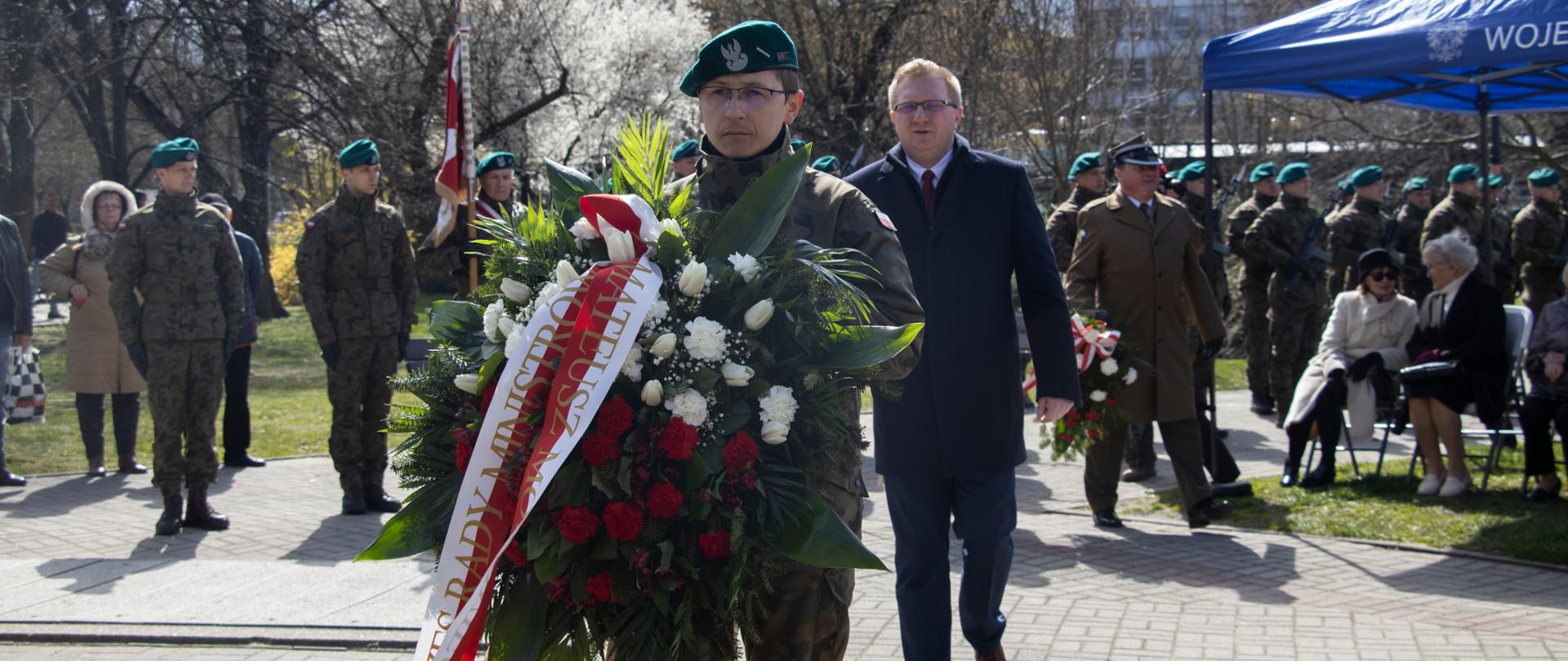 Na zdjęciu żołnierz trzymający wieniec z napisem Prezes Rady Ministrów. Za nim idący w kierunku pomnika wicewojewoda opolski. W tle pozostali uczestnicy uroczystości. 