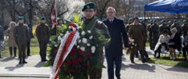 Na zdjęciu żołnierz trzymający wieniec z napisem Prezes Rady Ministrów. Za nim idący w kierunku pomnika wicewojewoda opolski. W tle pozostali uczestnicy uroczystości. 