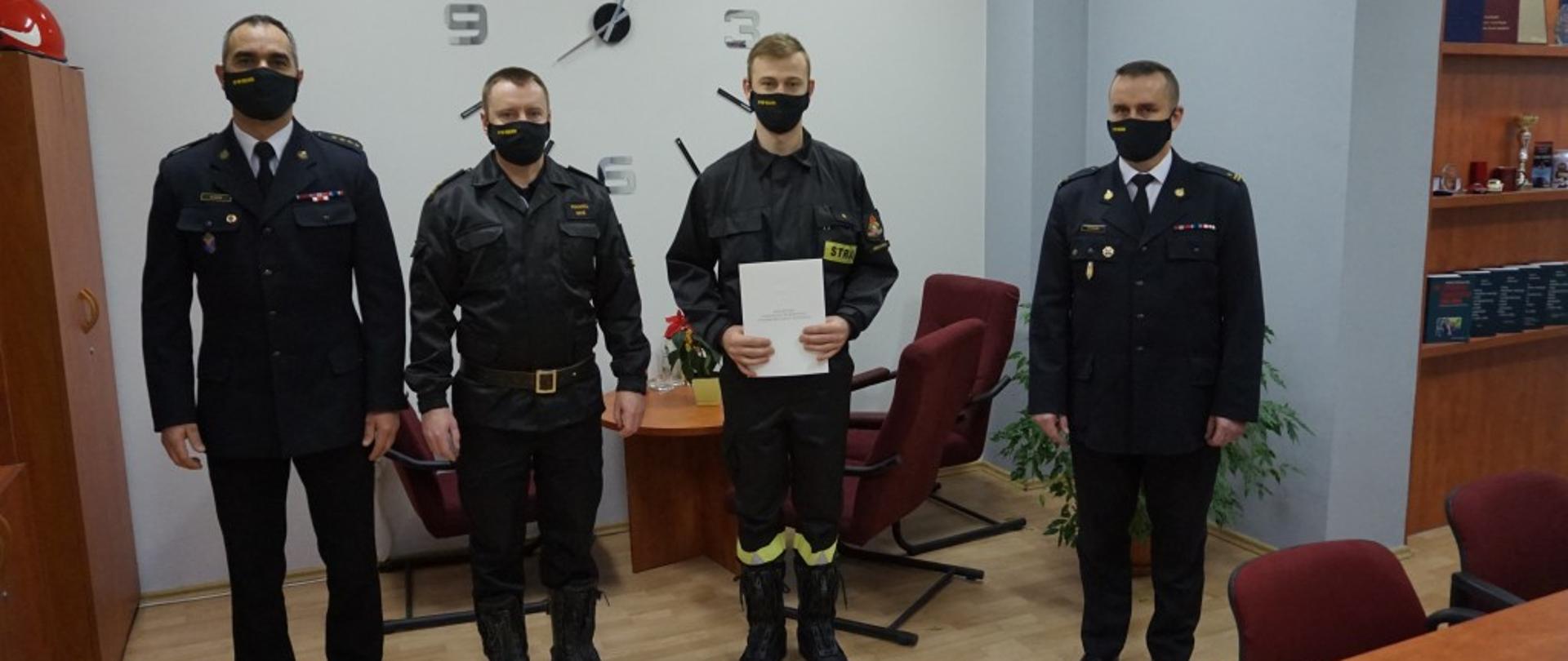 Na zdjęciu cztery osoby: Zastępca Komendanta Powiatowego st. kpt. Mariusz Idzik, Dowódca Jednostki Ratowniczo Gaśniczej mł. bryg. Paweł Miś, Awansowany asp. Grzegorz Kucharz, Komendant Powiatowy mł. brug. Michał Majda. Zdjęcie w gabinecie komendanta. 