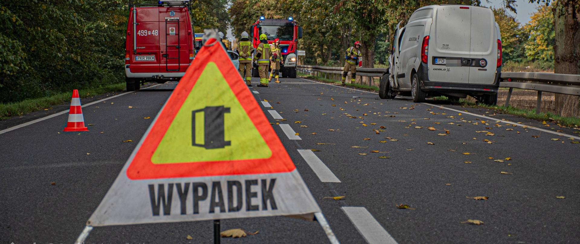 Zdjęcie na pierwszym planie przedstawia znak ostrzegawczy z napisem WYPADEK, oraz samochody straży pożarnej, oraz pojazd biorący udział w zdarzeniu.