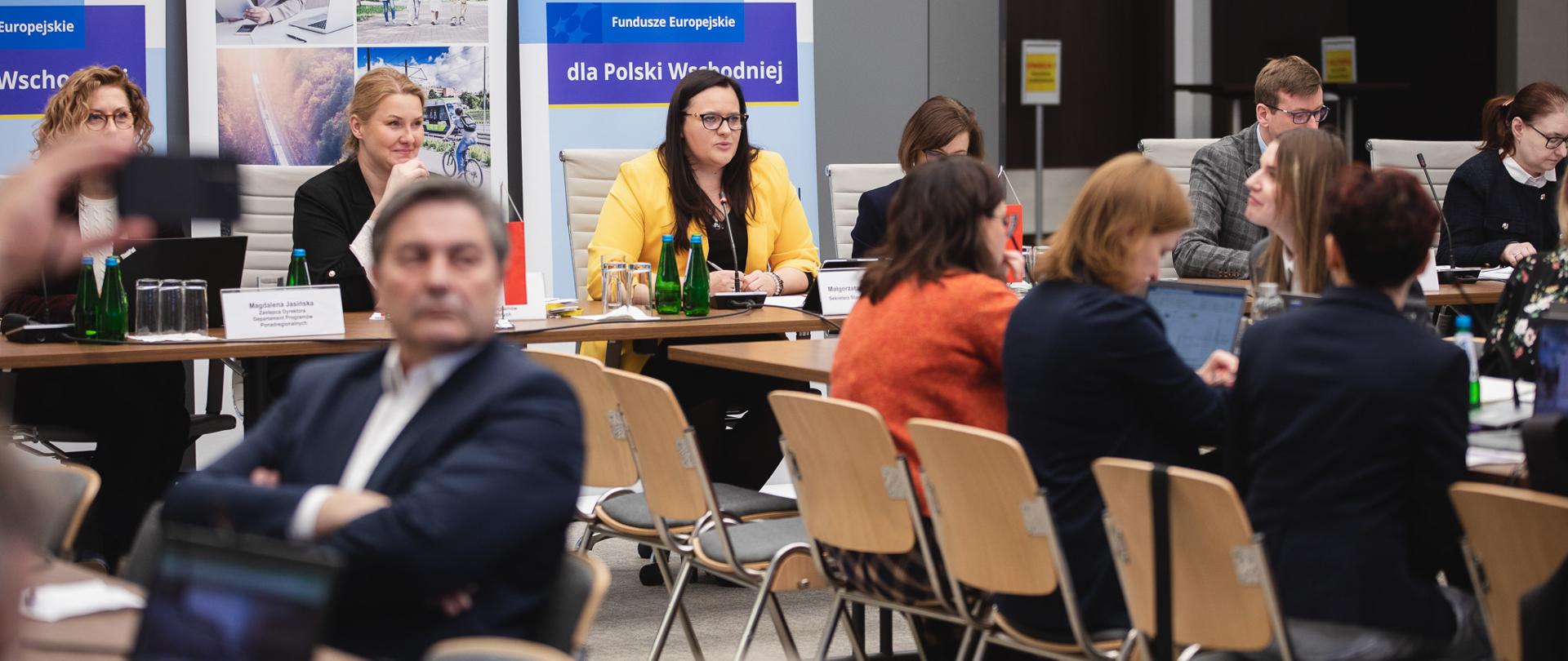 Grupa osób siedzi przy stołach w sali konferencyjnej. Na tle banerów wiceminister Małgorzata Jarosińska-Jedynak.