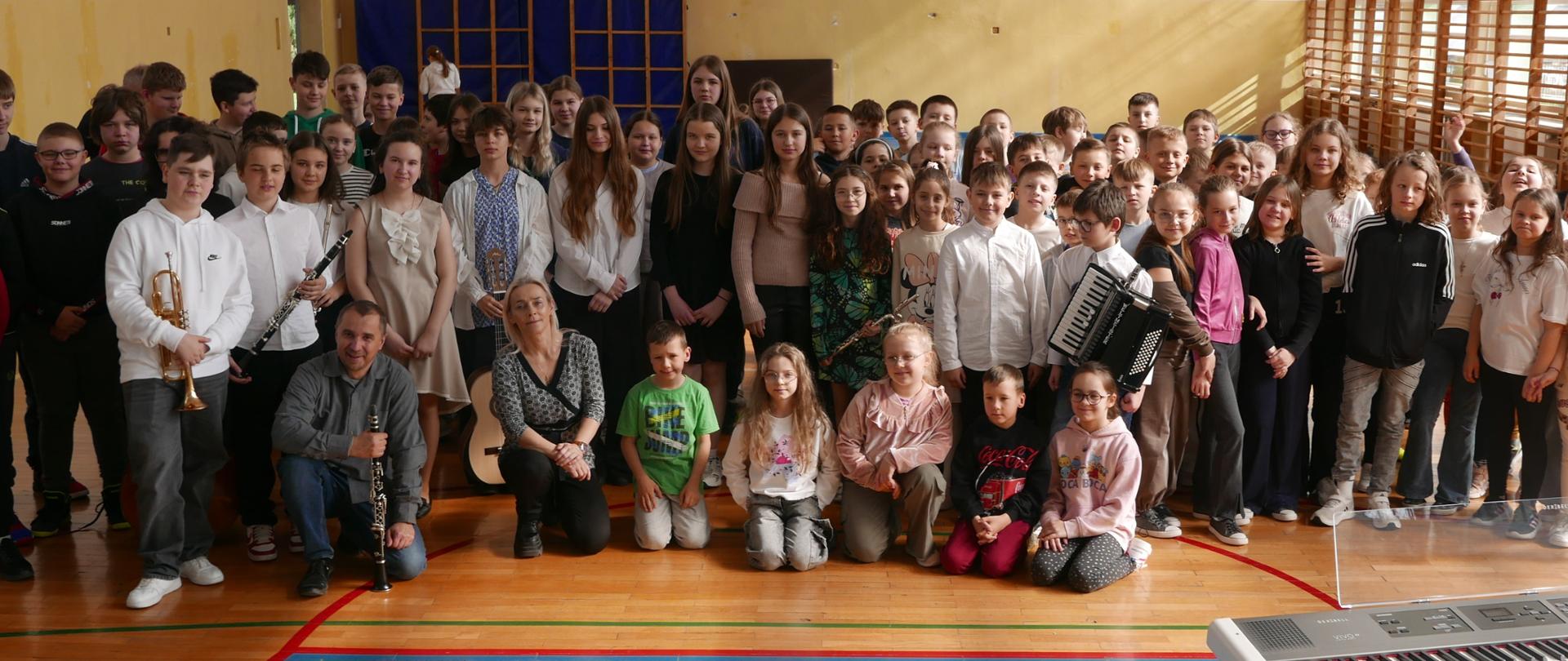 Dzieci i młodzież szkolna na wspólnej fotografii, niektórzy mają instrumenty muzyczne jak trąbka, klarnet, akordeon, flet poprzeczny. Po prawej stronie zdjęcia widzimy pianino cyfrowe z przezroczystym pulpitem nutowym. W tle widoczne są drabinki gimnastyczne.