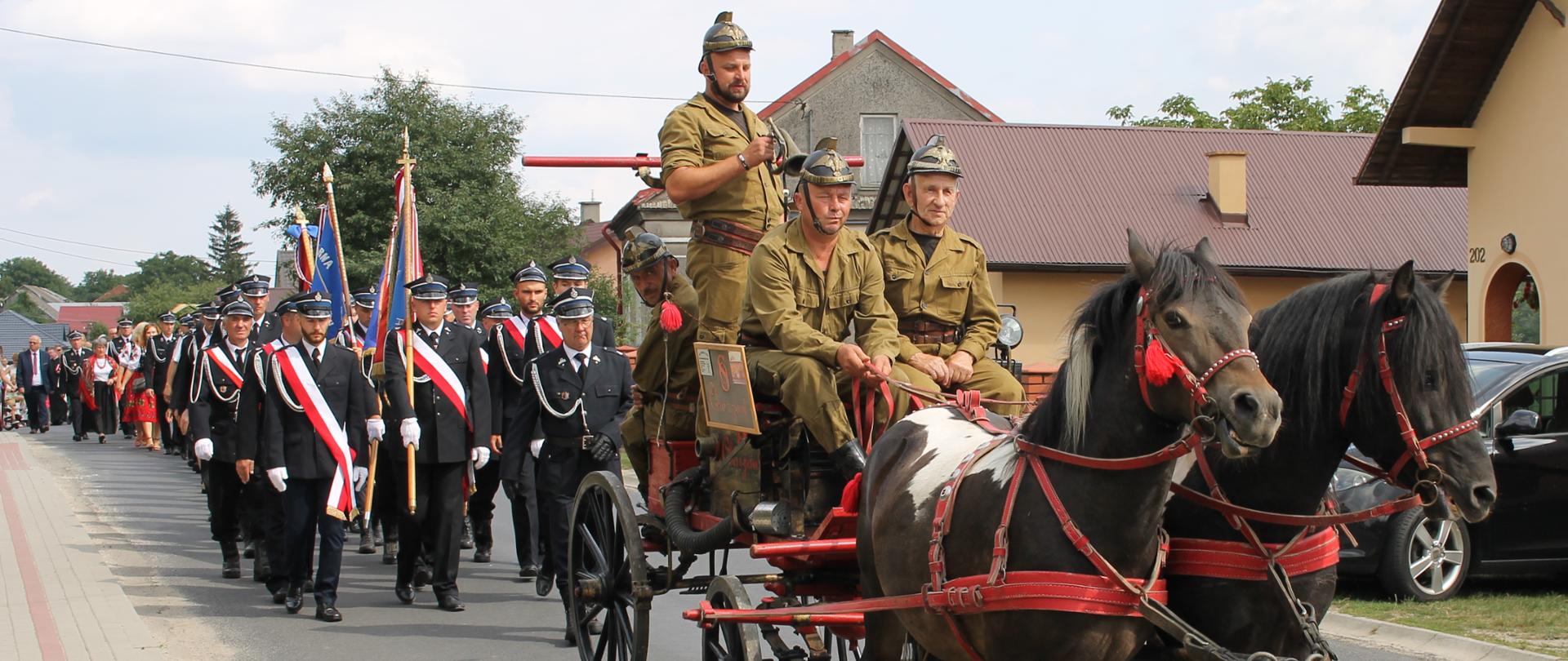 Na zdjęciu na pierwszym planie widzimy pożarniczą sikawkę, którą ciągną 2 konie. Obsadę sikawki stanowi czterech strażaków ubranych w tradycyjne umundurowanie strażackie. Na drugim planie widoczne są poczty sztandarowe, za nimi pozostałe osoby uczestniczące w przemarszu.