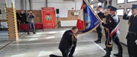 Fotografia przedstawia strażaka klęczącego przed sztandarem Państwowej Straży Pożarnej, który całuje jego róg na znak objęcia obowiązków komendanta powiatowego.