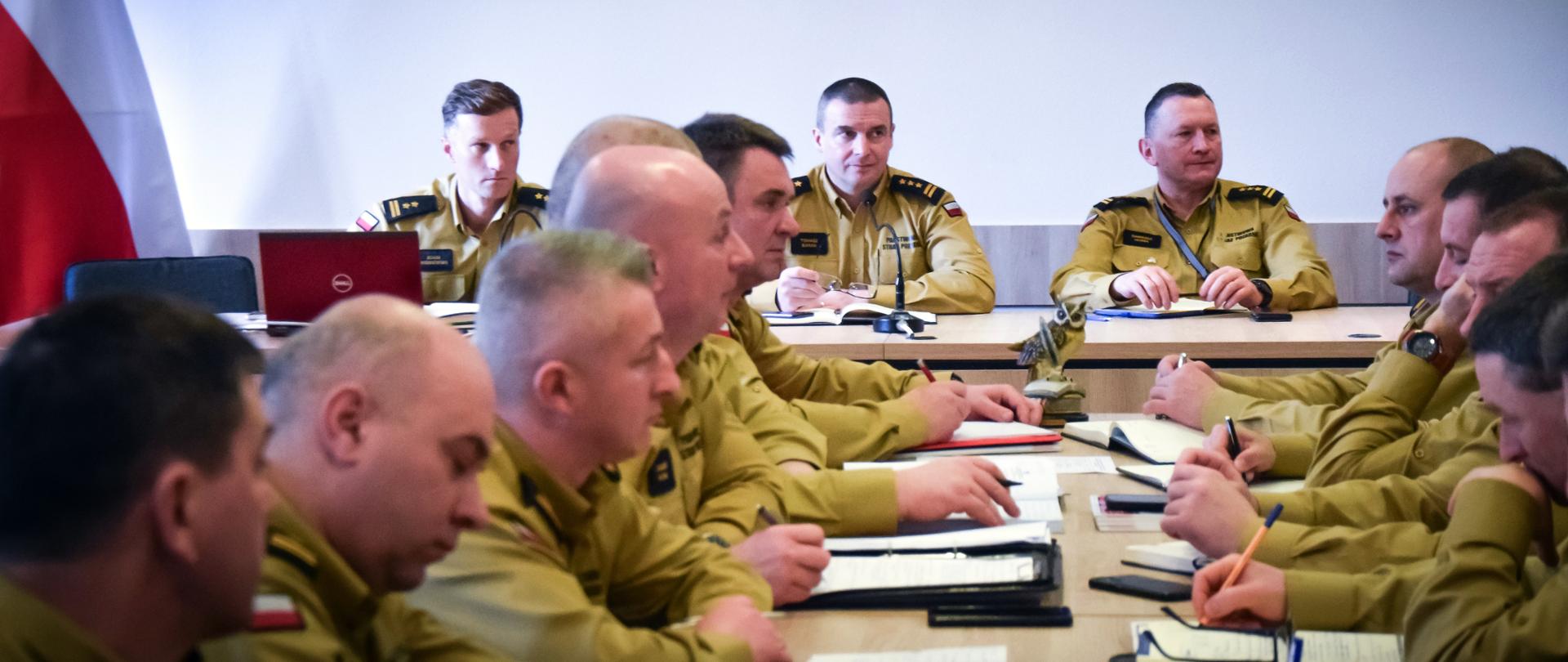 Zdjęcie zrobione w pomieszczeniu, podczas narady służbowej kadry kierowniczej Państwowej Straży Pożarnej. Na zdjęciu oficerowi PSP w piaskowych mundurach służbowych.