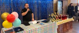 Mężczyzna przemawia do uczestników przez mikrofon, stojąc za stołem w udekorowanej sali. Na stole znajdują się dokumenty, laptop, kolorowe balony oraz liczne puchary i nagrody przygotowane dla uczestników turnieju. W tle widoczna jest błyszcząca dekoracja, a z boku stoją obserwujący wydarzenie uczestnicy.
