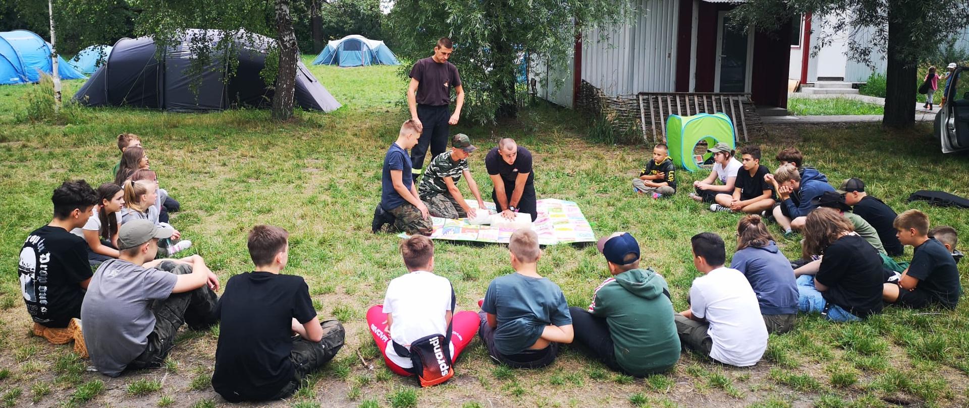 Strażacy pokazują pierwszą pomoc. Harcerze siedzą w półokręgu na trawie.