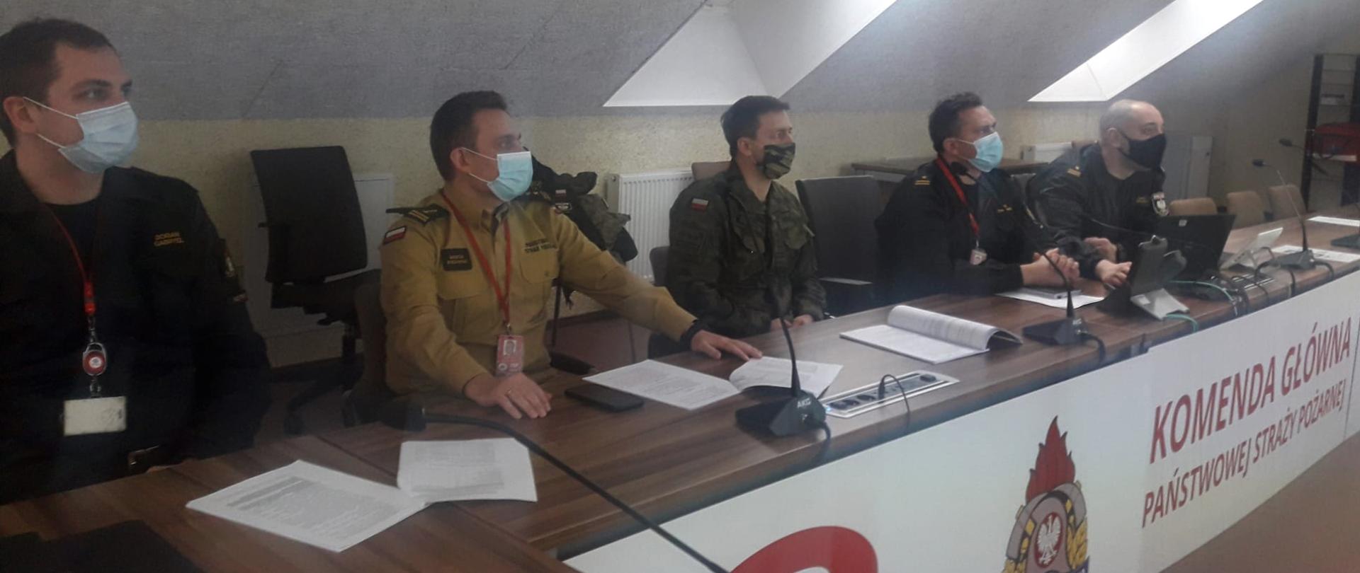 Czterech funkcjonariuszy PSP oraz jeden z wojskowej ochrony przeciwpożarowej (po środku) siedzą przy biurku podczas spotkania, patrzą przed siebie