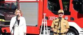 Kobieta w białym stroju oraz strażak w mundurze w kolorze musztardowym stoją przed mikrofonami za nimi stoi sztaluga oraz samochody pożarnicze.