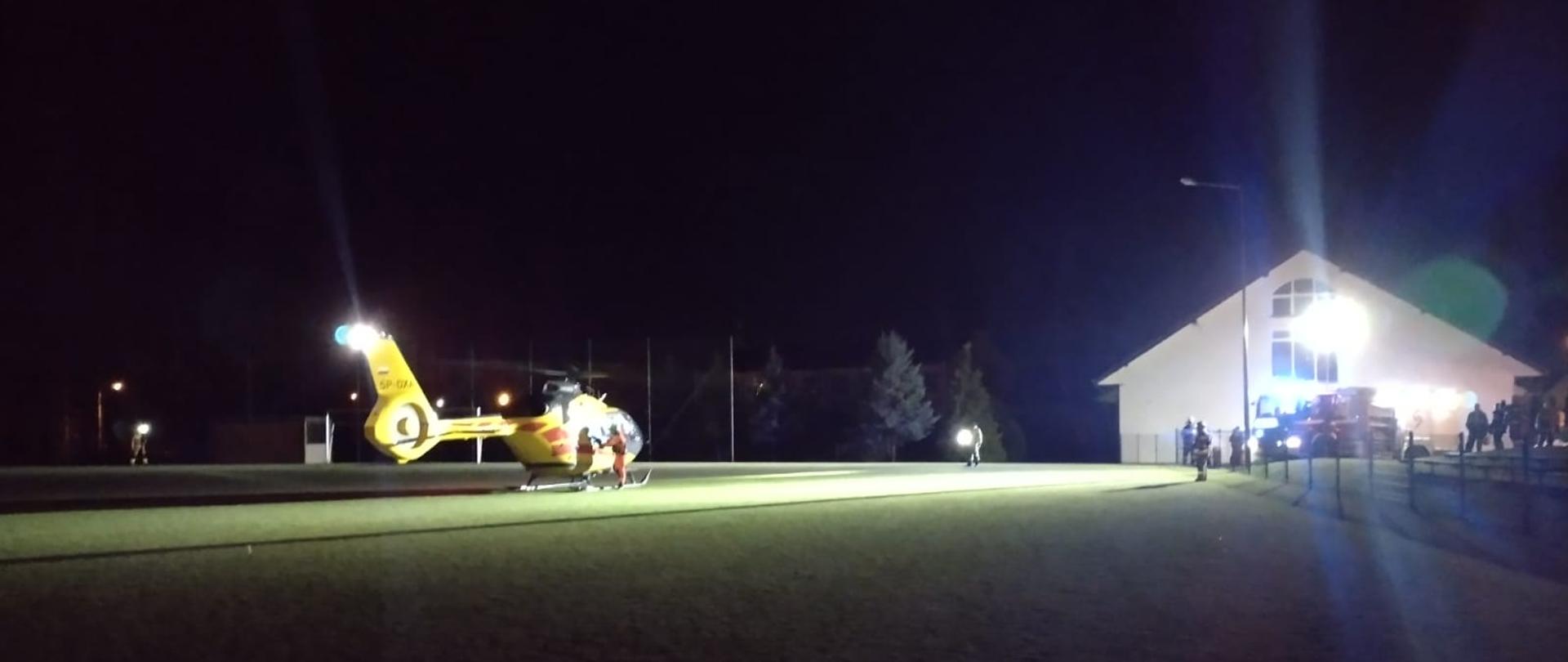 Na zdjęciu żółty śmigłowiec LPR. Ma włączone światło. Miejsce to boisko sportowe z zieloną trawą. Boisko ogrodzone siatką. Pora nocna. Po prawej samochody straży pożarnej z włączonymi światłami mijania.