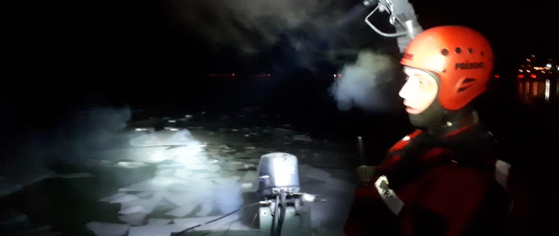 Strażak z Woprowcem płyną łodzią na jeziorze na którym znajduje się kra lodowa. Teren oświetlają latarkami.