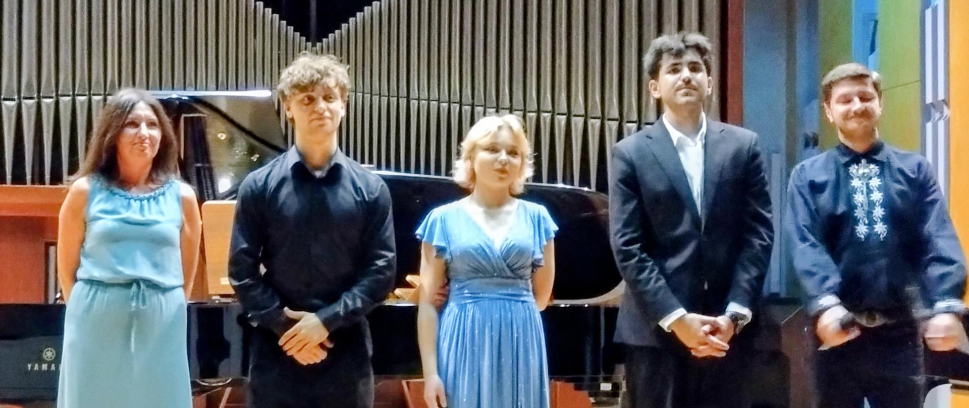 Zdjęcie przedstawia pięcioro elegancko ubranych artystów (dwie kobiety i trzech mężczyzn) stojących w rzędzie na scenie sali koncertowej lub filharmonii. Postacie: Od lewej stoi kobieta w jasnoniebieskiej, długiej sukni, obok niej młody mężczyzna w czarnej koszuli i spodniach, następnie blondynka w niebieskiej, zwiewnej sukni, mężczyzna w ciemnym garniturze oraz mężczyzna w ciemnej koszuli ze zdobieniami/haftem na piersi. Tło: Za artystami widać duży fortepian (marki Yamaha) oraz imponujące piszczałki organów zajmujące centralną część ściany. Klimat: Zdjęcie wygląda na moment ukłonów lub podziękowań po zakończonym występie muzycznym.