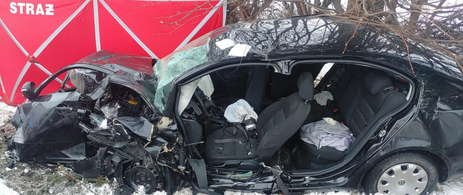 Rozbity pojazd marko Skoda, widziany z jego prawej strony, za nim po lewej stronie
stoi rozstawiony czerwony parawan z napisem straż. Pojazd znajduje się poza drogą. W tle krzaki i zarośla.