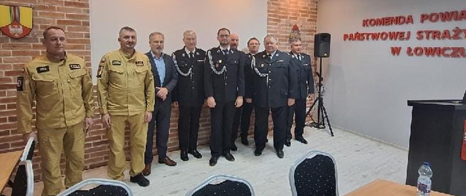 Na zdjęciu widać dziewięciu mężczyzn pozujących do zdjęcia. Dwóch ubranych jest w żółte stroje piaskowe, jeden w garnitur a reszta w ciemne stroje wyjściowe strażackie ochotniczych straży pożarnych. Zdjęcie zrobione jest wewnątrz budynku. Na ścianie widać część napisu, jednak można się domyśleć, że jest napisane Komenda Powiatowa Państwowej Straży Pożarnej w Łowiczu. Na pierwszym planie widać krzesła stół, na którym leżą dokumenty oraz talerz z ciastkami. Jest zapalone światło.