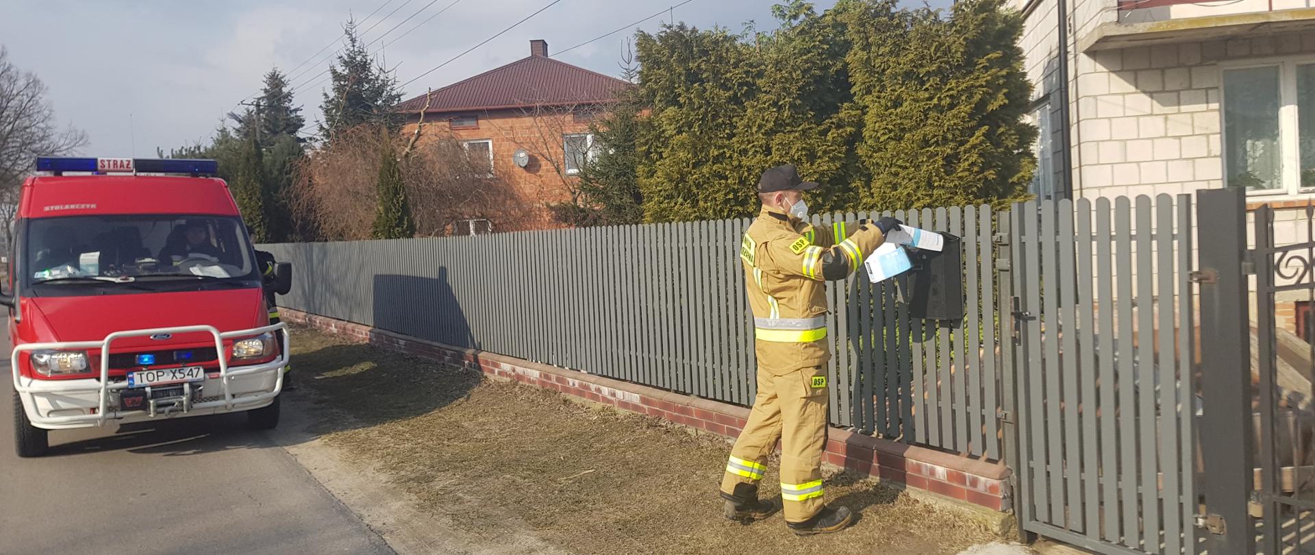 Zdjęcie przedstawia strażaka OSP wrzucającego ulotkę zawierającą informację o szczepieniach do skrzynki pocztowej. W tle widać zabudowania oraz czerwony samochód OSP.