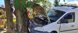 Biały pojazd osobowy VW Caddy, przodem uderzył w drzewo. W tle widoczni są strażacy oraz ratownicy medyczni.