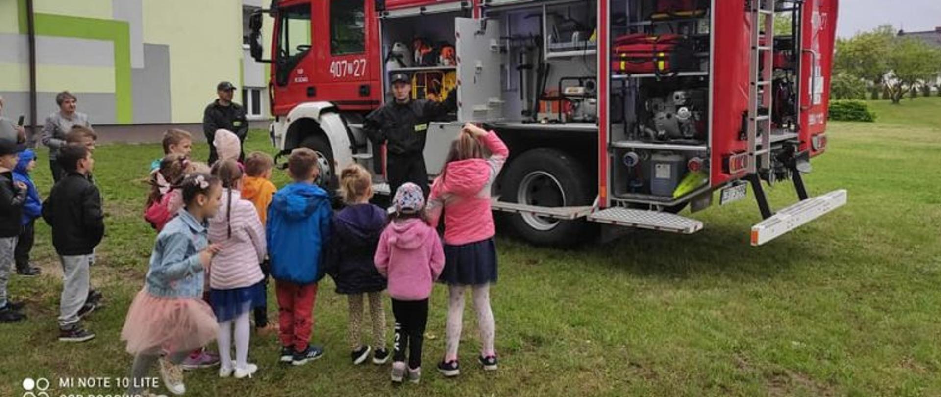 Zdjęcie wykonane przed szkołą. Centralny punkt zdjęcia stanowi wóz gaśniczy i stojący przy nim strażak, który demonstruje sprzęt. Przed wozem stoją dzieci i słuchają strażaka.