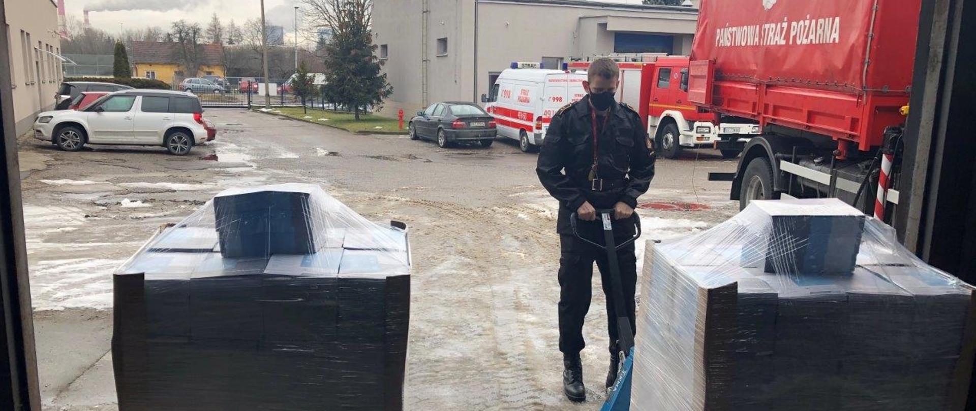 Parking między budynkami. Samochody Państwowej Straży Pożarnej. Dwie palety ulotek obok strażak.