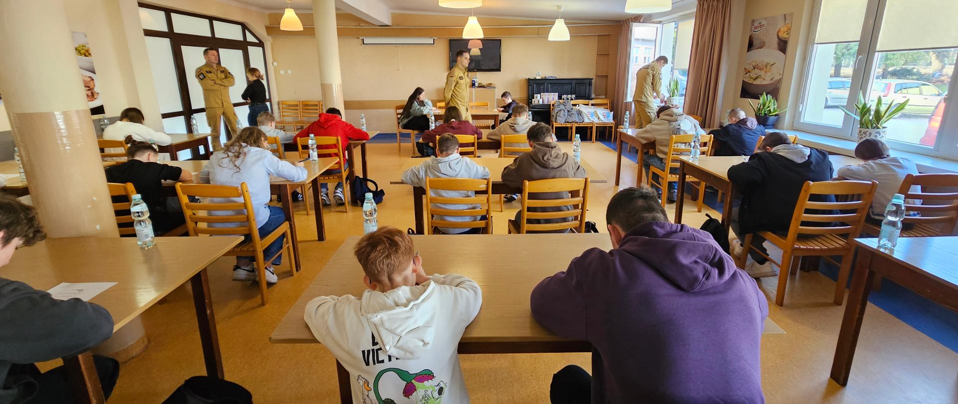 Zdjęcie przedstawia jasne, przestronne pomieszczenie, przypominające świetlicę lub stołówkę, w którym grupa młodzieży siedzi przy drewnianych stolikach i wypełnia arkusze papieru.
Uczestnicy: Około dwudziestu młodych osób siedzi tyłem lub bokiem do obiektywu, pochylając się nad zadaniami. Na stołach widoczne są butelki z wodą mineralną.
Osoby nadzorujące: W głębi sali oraz przy oknie stoją trzej mężczyźni w piaskowych mundurach służbowych (prawdopodobnie strażacy lub przedstawiciele innej służby mundurowej), którzy czuwają nad przebiegiem wydarzenia.
Wnętrze: Sala ma żółte ściany i duże okna po prawej stronie, przez które wpada dzienne światło. Pod sufitem wiszą liczne białe lampy w kształcie dzwonów. W tle widać czarny instrument (pianino lub fortepian cyfrowy) oraz ekran projekcyjny.
Atmosfera: Scena sugeruje trwanie konkursu wiedzy lub warsztatów edukacyjnych w spokojnej, skupionej atmosferze.
