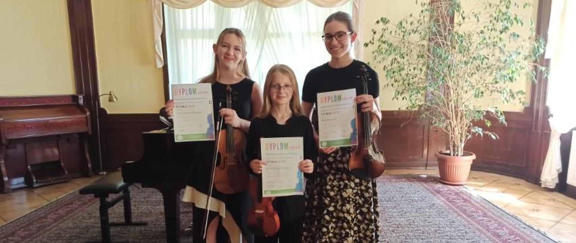 Zdjęcie kolorowe. Trzy uczennice trzymają w rękach skrzypce i dyplomy. W tle fortepian, stołek do fortepianu, okno z kremowymi zasłonami. Z lewej strony fisharmonia, z prawej strony rozłożysty figowiec benjamina.