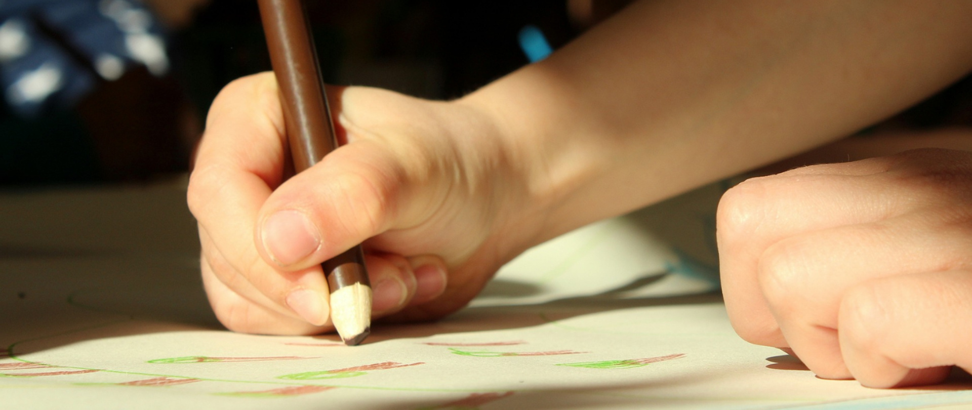 Grafika przedstawia dłonie dziecka trzymającego kredkę i rysującego obrazek.