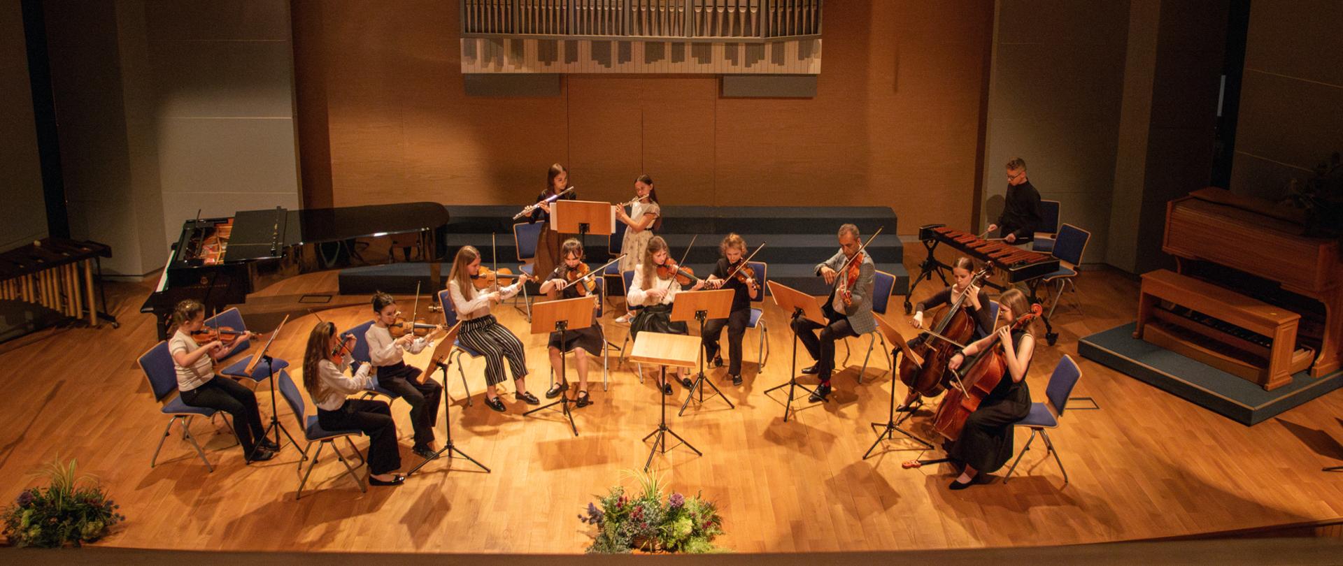 Obraz przedstawia widok z góry na młodą orkiestrę grającą na scenie w sali koncertowej. Scena ma drewnianą podłogę, a w tle znajduje się duży prospekt organowy na ścianie oraz reflektory sceniczne. W kilku rzędach siedzą muzycy, grający na różnych instrumentach, głównie smyczkowych takich jak skrzypce i wiolonczele, ale także na fletach. Po lewej stronie sceny widoczny jest fortepian, a po prawej marimba. Na pierwszym planie, w dolnej części obrazu, widoczne są dwie kompozycje kwiatowe. Oświetlenie sceny jest jasne, skupiające się na muzykach i instrumentach.