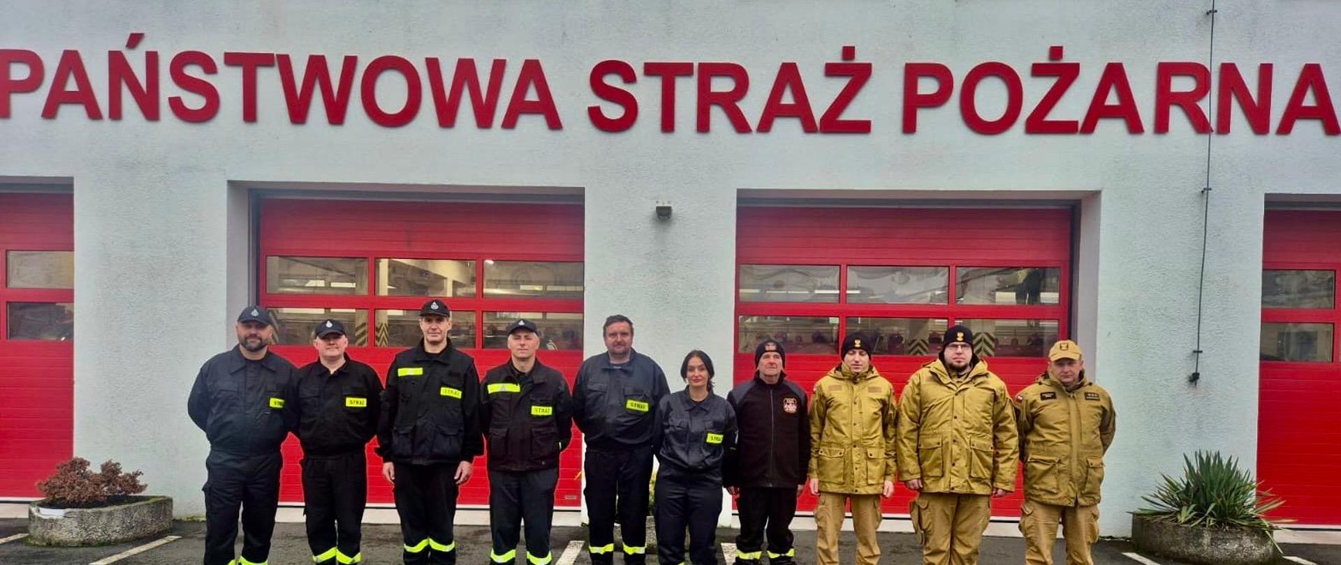 Na tle czerwonych bram strażnicy stoi zespół ludzi w jednakowym szyku. Różnorodność umundurowania i postaw pokazuje jedność mimo odmiennych ról. To moment zatrzymany między codzienną służbą a wspólną odpowiedzialnością — spokojny, ale pełen gotowości. Budynek za nimi nie jest tylko zapleczem technicznym, lecz symbolem miejsca, z którego wyrusza pomoc.
