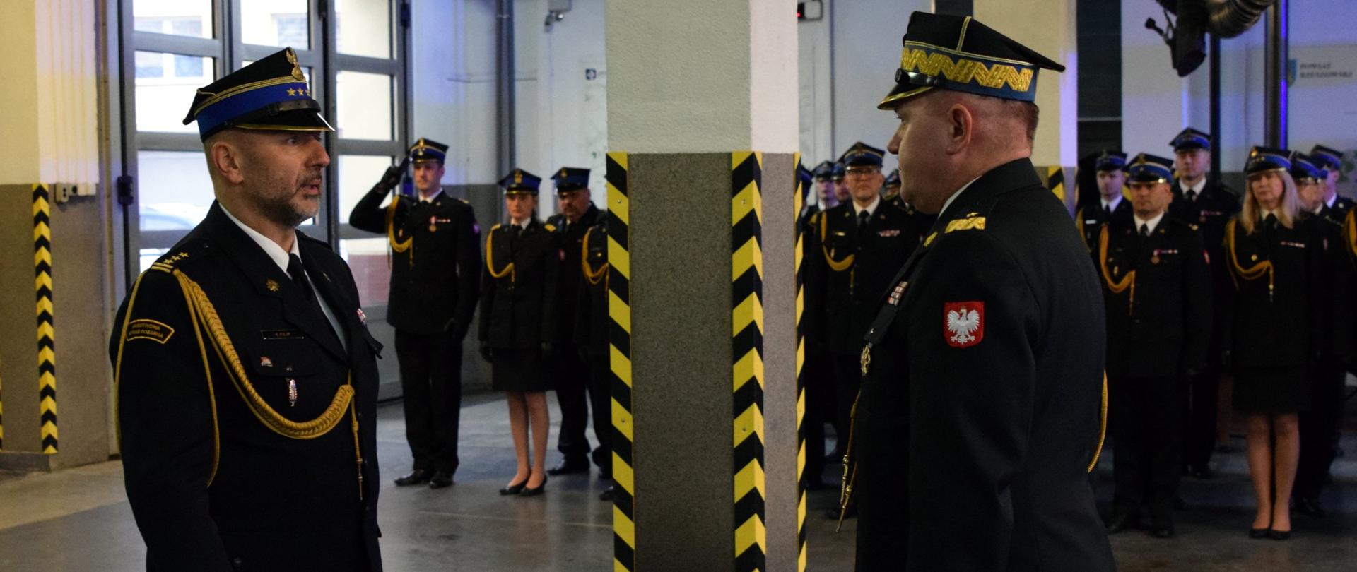 Zdjęcie przedstawia dwóch strażaków stojących naprzeciwko siebie. W tle widoczny jest pododdział strażaków.