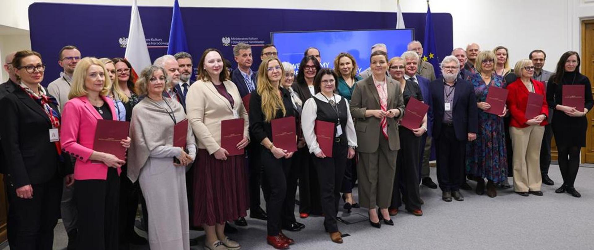 Fotografia grupowa kilkudziesięciu osób pozujących na tle ścianki Ministerstwa Kultury i Dziedzictwa Narodowego. Wiele osób trzyma w dłoniach bordowe teczki z dyplomami. W tle stoją flagi Polski i Unii Europejskiej.