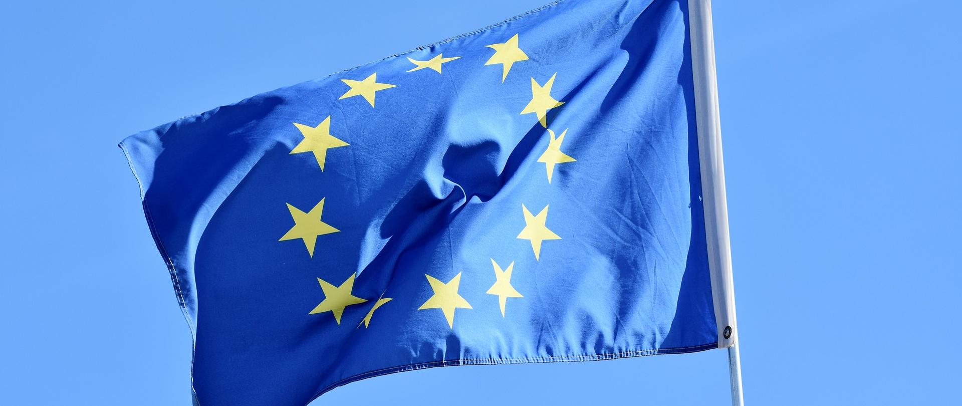 Na tle nieba zawieszona na maszcie flaga Unii Europejskiej - okrąg złożony z dwunastu złotych gwiazd na błękitnym tle