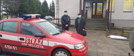 Na drodze przy chodniku stoi czarowny samochód osobowy z białym napisem Straż OSP Wilczęta. Przy aucie na chodniku stoi strażak w mundurze i maseczce obok starszy pan w czarnym płaszczu z czarną teczka w czapce i maseczce.
W tle na zdjęciu budynek gminnego ośrodka zdrowia, wejście do budynku po schodach, duże przeszklone drzwi. Po lewej stronie drzwi napis NFZ.
