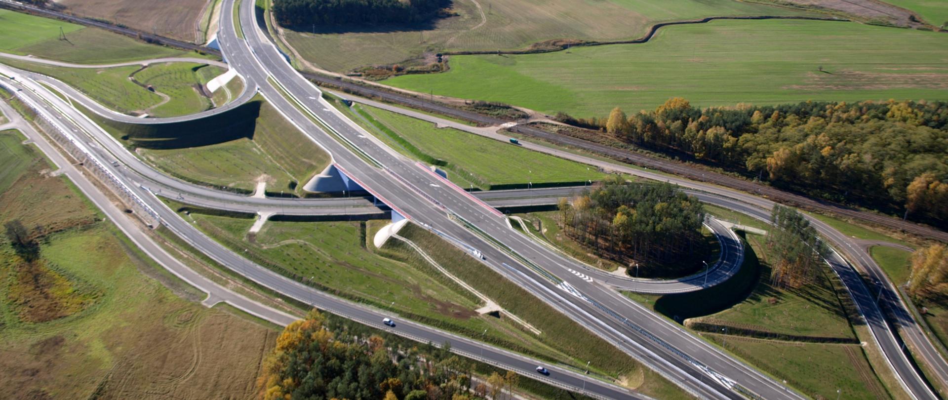 Zdjęcie przedstawiające węzeł drogowy Poznań Północ na drodze ekspresowej S11. Węzeł widoczny z powietrza przebiegający między łąkami, polami i zabudową magazynową. Na jednej z łącznic doprowadzających ruch do węzła widoczne poruszające się pojazdy.