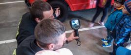
Zdjęcie przedstawia strażaka i dzieci podczas prezentacji kamery termowizyjnej na tle pojazdu trażackiego koloru czerwonego.
