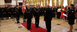 Na czerwonym dywanie stoi dwóch strażaków, trzeci przed nimi, zwrócony przodem do tamtych. Za nimi trzech innych ze sztandarem. W dai widać kilkadziesiśt osób, gości uroczystości. Z prawej strony strażak kobieta, trzyma dokumenty.