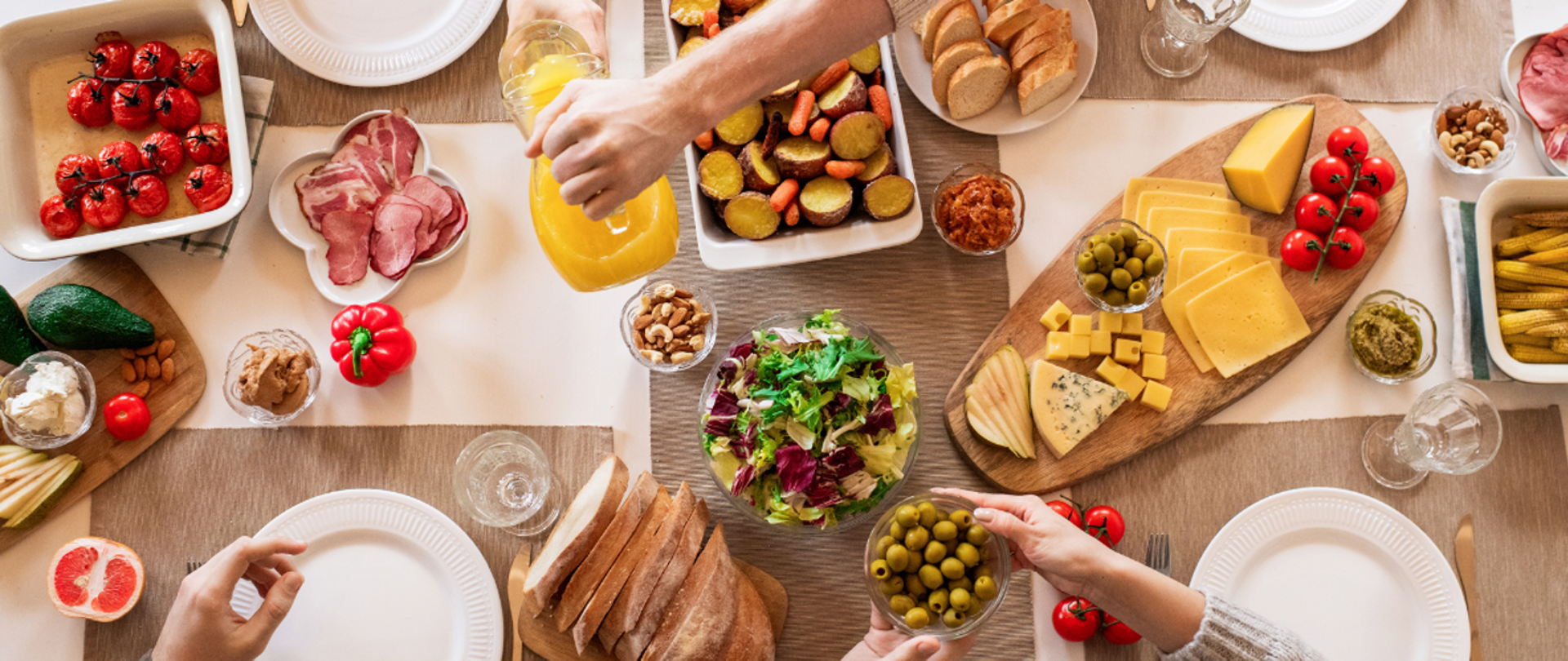 Zdjęcie przedstawiające stół zastawiony żywnością i talerzami. Na półmiskach i deskach do krojenia znajdują się warzywa, sery, wędliny, owoce i orzechy. Na zdjęciu widać dłonie sięgające po jedzenie.
