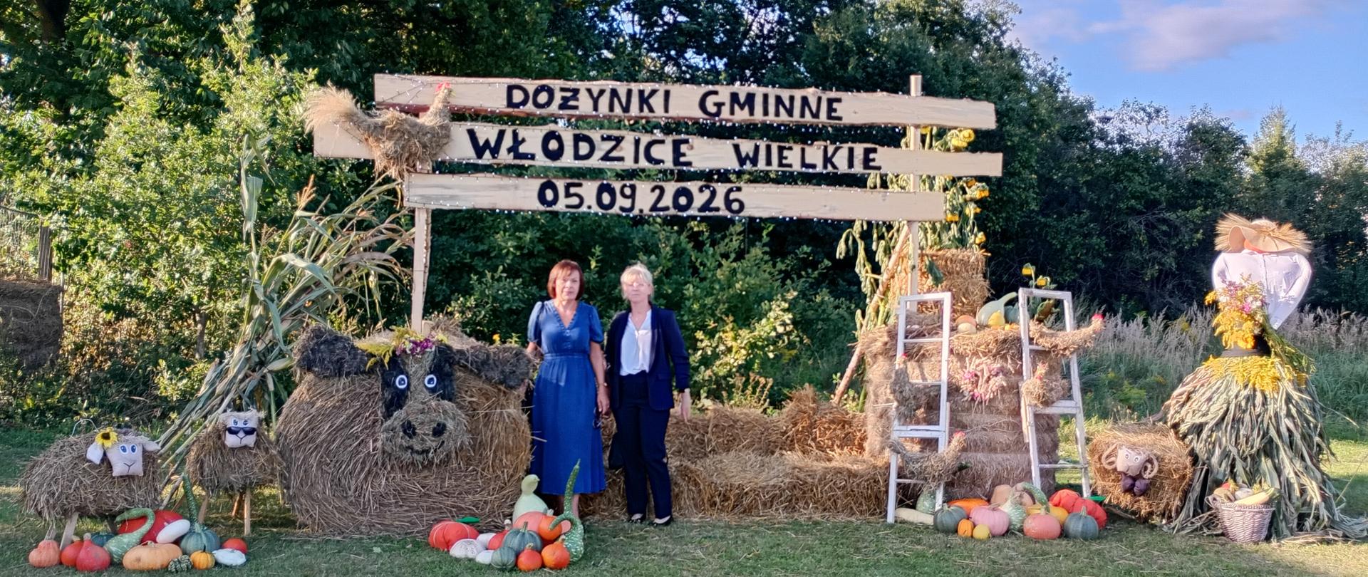 Dekoracja dożynkowa z napisem „Dożynki Gminne Włodzice Wielkie 05.09.2026”. Na środku stoją dwie kobiety w eleganckich strojach, obok siebie, na tle dekoracji ze słomy, warzyw i kwiatów. Po bokach znajdują się słomiane figury i ozdoby.