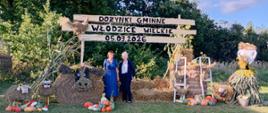 Dekoracja dożynkowa z napisem „Dożynki Gminne Włodzice Wielkie 05.09.2026”. Na środku stoją dwie kobiety w eleganckich strojach, obok siebie, na tle dekoracji ze słomy, warzyw i kwiatów. Po bokach znajdują się słomiane figury i ozdoby.