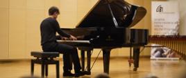 Zdjęcie przedstawia innego ucznia starszej klasy grającego na fortepianie na scenie sali koncertowej, w tle baner szkoły