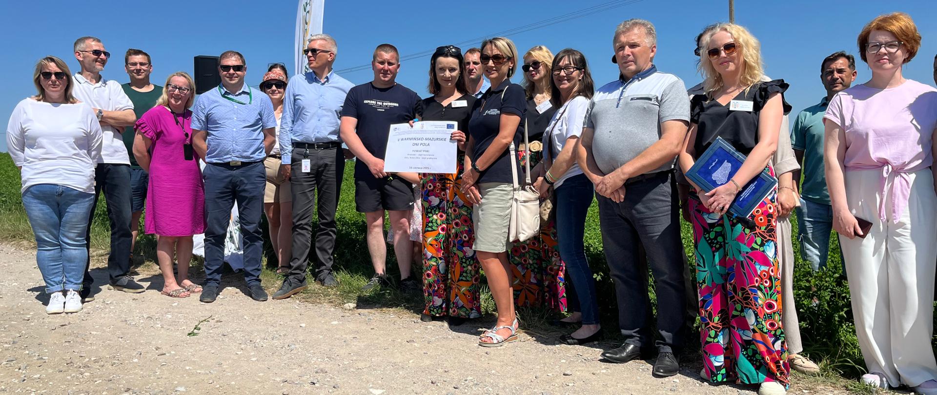 17 osób, kobiet i mężczyzn, 1 osoba trzyma plakat informacyjny.
W tle widoczna uprawa lucerny