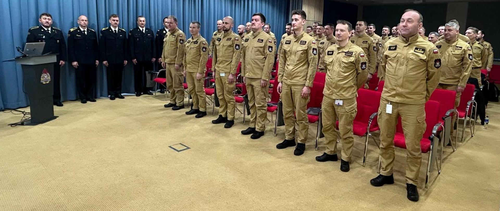 W Sali konferencyjnej z niebieską kotarą i czerwonymi krzesłami stoją strażacy PSP w piaskowych mundurach służbowych, a stojący pod ścianą w czarnych mundurach wyjściowych. 