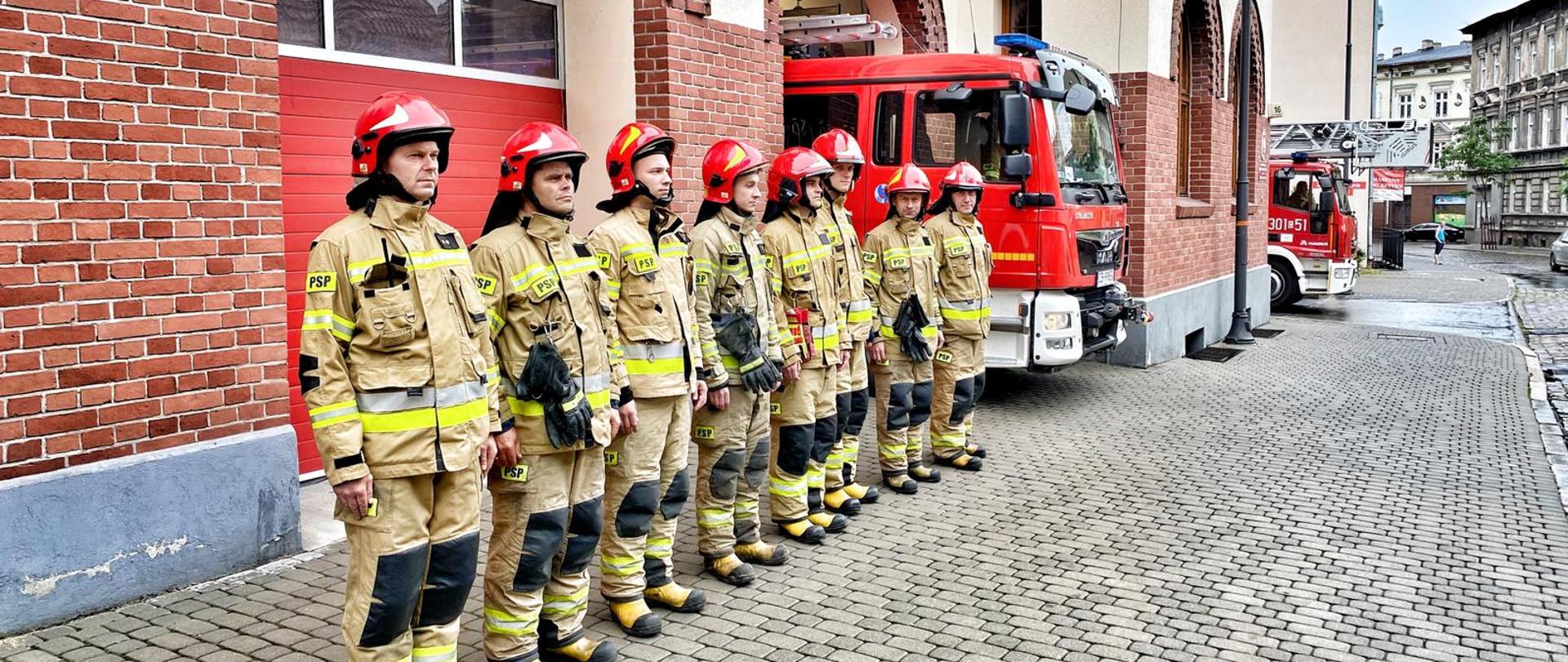 Strażacy stojący w szeregu w żółtych ubraniach specjalnych przed budynkiem Jednostki Ratowniczo-Gaśniczej nr 1 w Bydgoszczy, w tle elewacja frontowa budynku i widok częściowo wystawionych dwóch wozów pożarniczych.