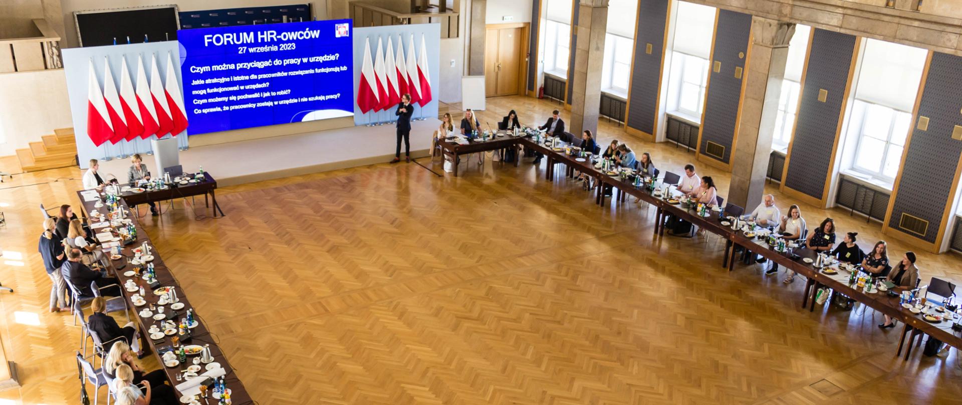 W dużej sali, przy dwóch długich stołach siedzi kilkadziesiąt osób. Na środku wyświetlana prezentacja, po dwóch stronach biało-czerwone flagi.