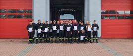 grupa dwudziestu strażaków ratowników stoi przed otwartą bramą wyjazdową jednostki ratowniczo-gaśniczej w łowiczu, w tle wewnątrz garażu auto strażackie, ratownicy stojąpozując do zdjęcia trzymają w ręku certyfikaty ukończenia kursu dla strażaków ratowników ochotniczych straży pożarnych, ubrani są w czarne ubranie koszarowe z żółtymi pasami na nogawkach, maja na nogach wysokie buty z cholewką, ośmiu z nich ma na głowie czapkę z daszkiem barwy czarnej, w tle po lewej i prawej stronie dwie zamknięte bramy wyjazdowe barwy czerwonej, teren przed garażem pokryty kostką brukową