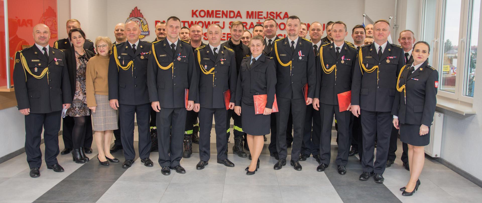 Na zdjęciu strażacy w mundurach koloru czarnego na tle ściany z napisem i flagami.