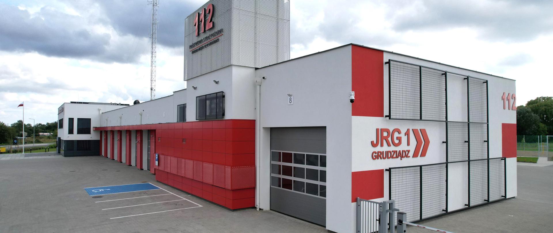 Szaro – czerwony budynek komendy i jednostki strażackiej z siwymi bramami wyjazdowymi widziany od frontu z boku.