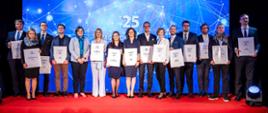 Zdjęcie zbiorowe, na przykrytym czerwonym dywanem podwyższeniu z przodu sali stoi grupa 15 osób, piąta od lewej wiceminister Zioło-Pużuk, wszyscy pokazują trzymane w rękach dyplomy, za nimi na ścianie ekran z napisem 25 na niebieskim tle.